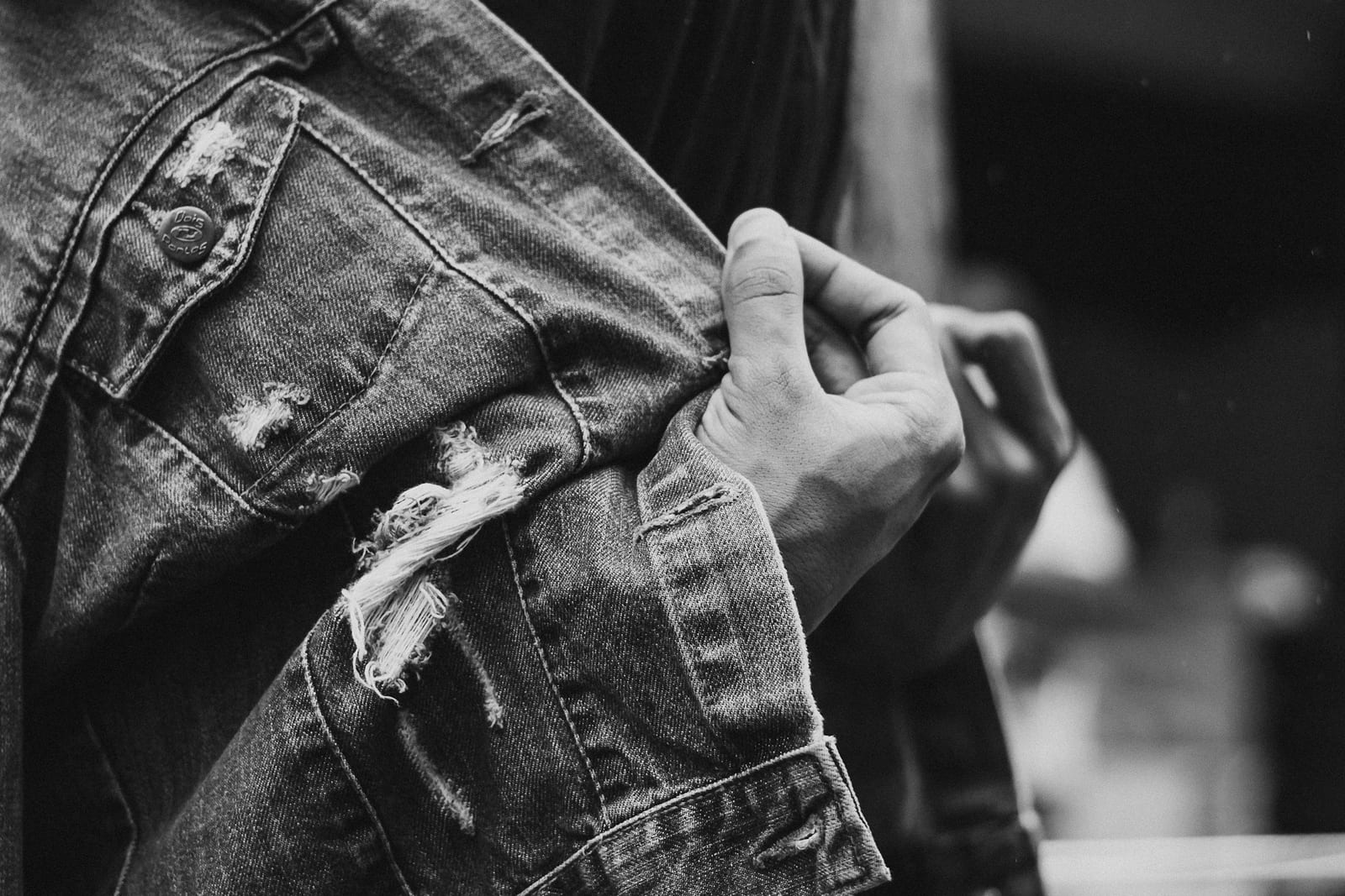 A black and white close-up of a denim jacket sleeve, worn through on the forearm with threads hanging from the tear, and the cuff edge beginning to fray