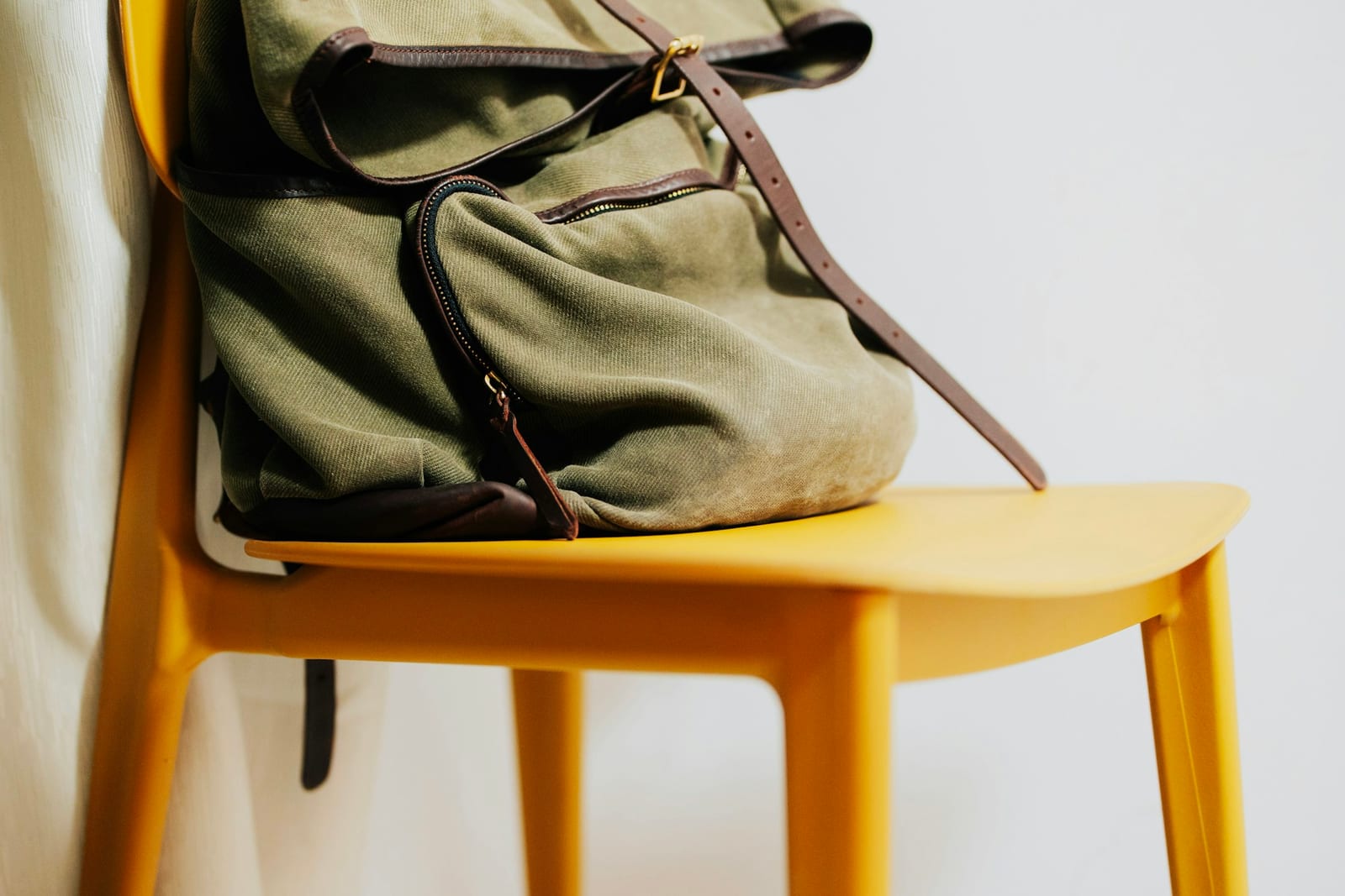 A green canvas rucksack with leather straps and a buckled flap, resting on a yellow chair, the canvas along its lower edge rubbed pale and dusty