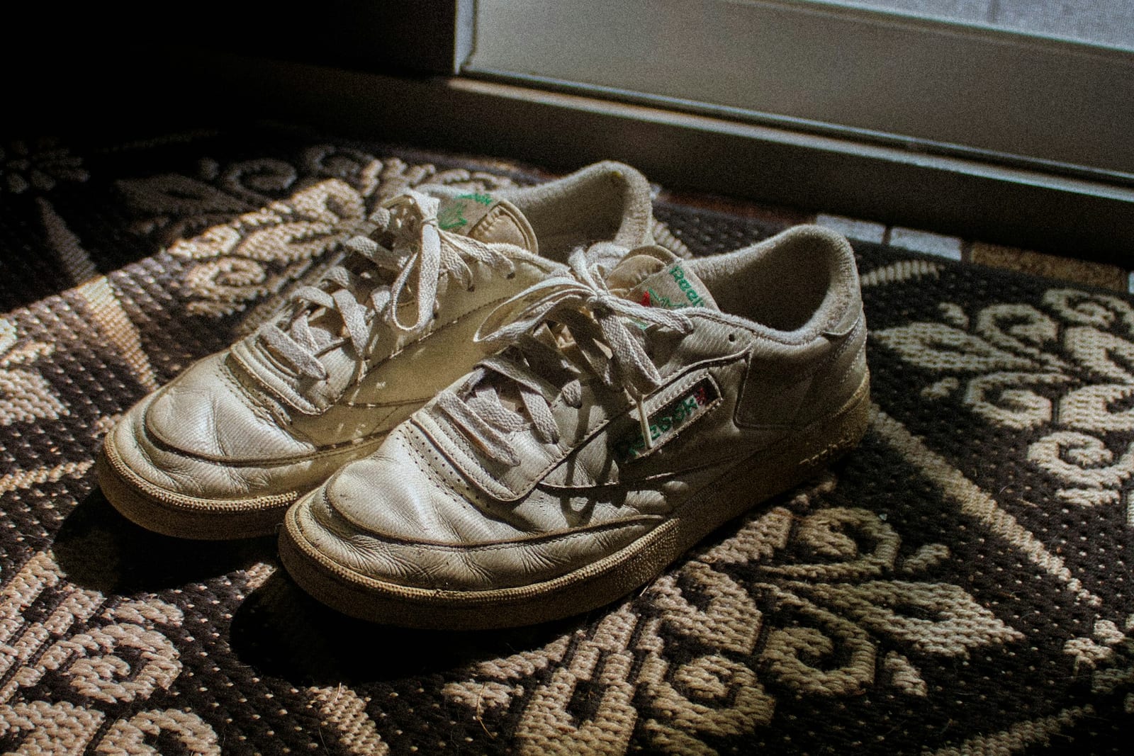 A pair of worn white leather trainers on a dark patterned doormat, creased across the toes, the soles grey and the leather scuffed