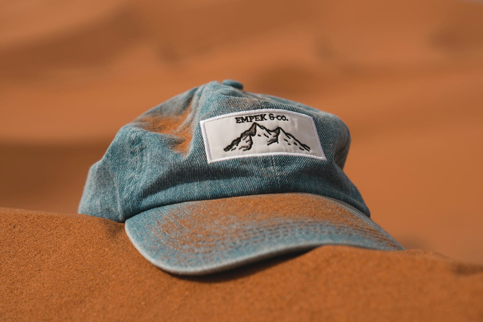 A blue cotton baseball cap resting in orange sand, the colour gone pale and powdery across the crown and peak, with fine dust worked into the weave