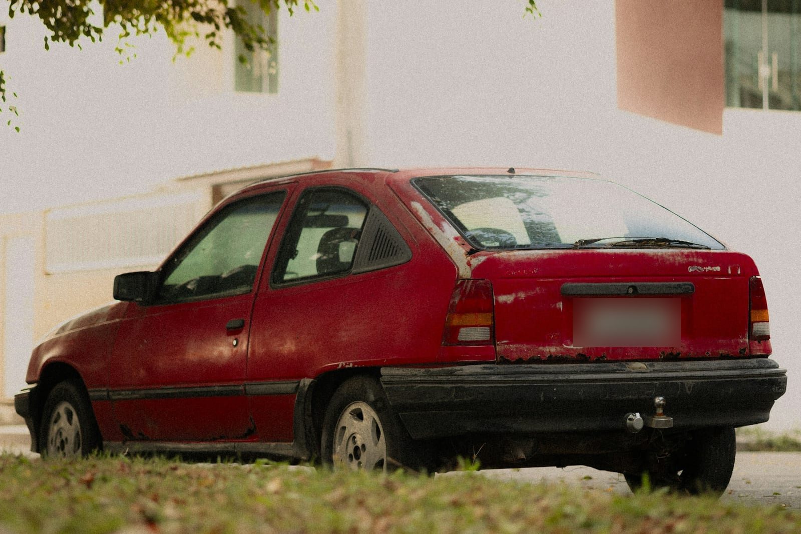 An old red hatchback parked on grass, its paint dull and chipped along the tailgate edge and bumper while the doors are still a deep red