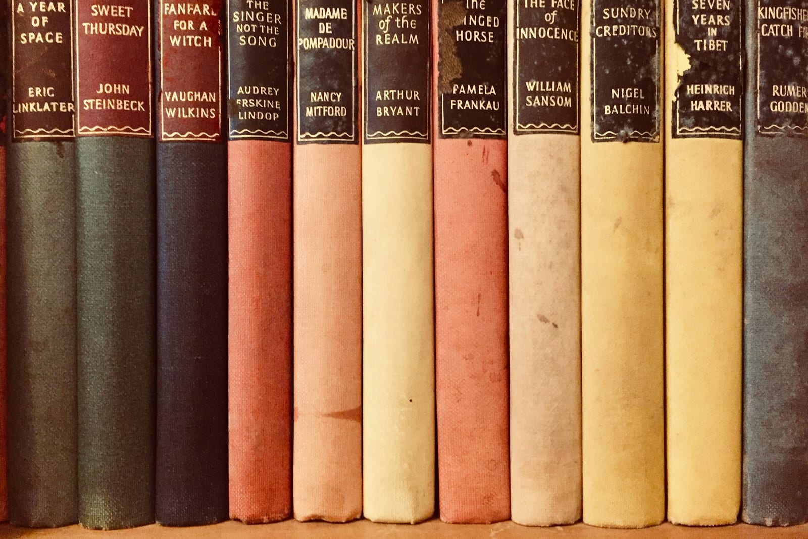 A row of old cloth-bound books standing together, their spines running from deep green, navy and red to pale pink, cream and chalky yellow, with the paper labels worn at the edges