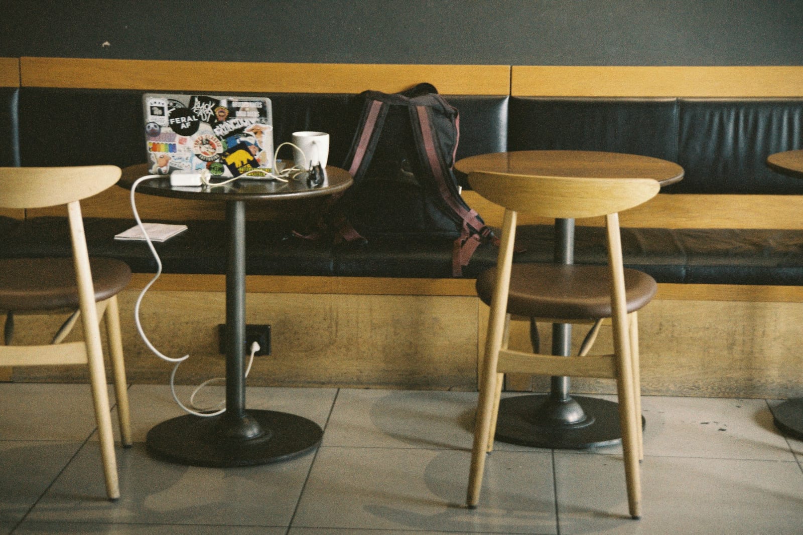 A café table just after its owner stepped away: a stickered laptop open and plugged into the wall socket by a white cable, a white mug beside it and a dark backpack left on the bench behind. The table next to it is bare