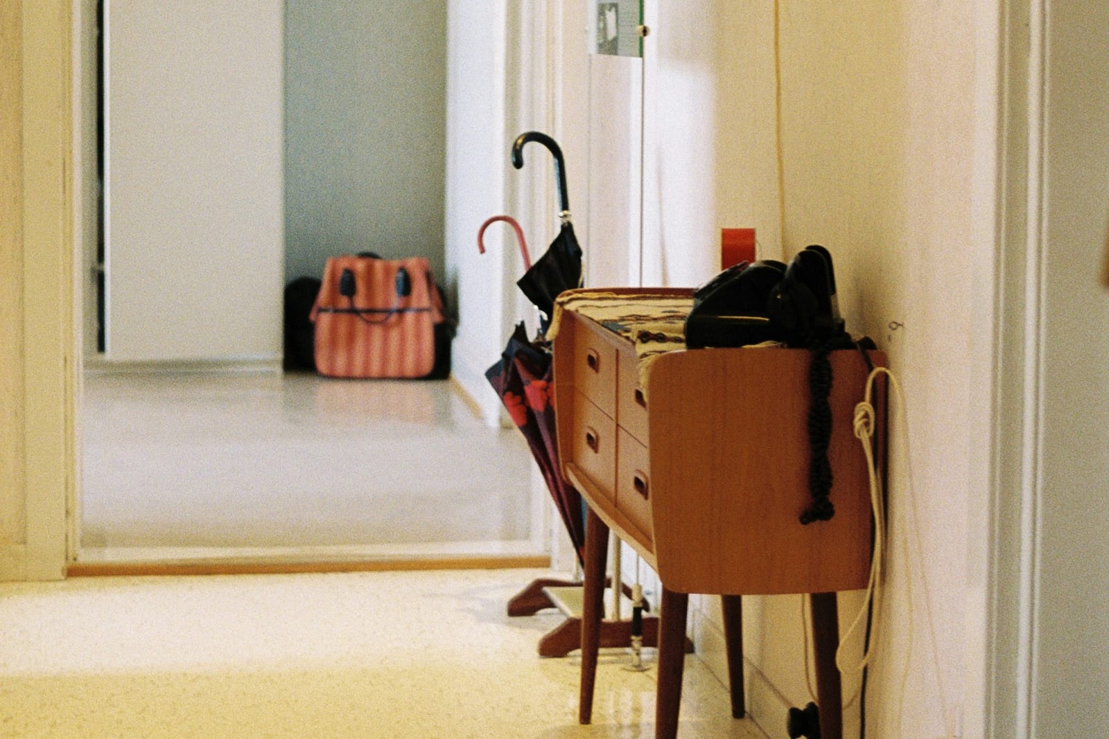 A bright, tidy hallway with a doorway at the far end. At the right-hand edge of the frame two umbrellas lean against a wooden chest of drawers with a dark bag on top, and a striped bag sits on the floor at the far end